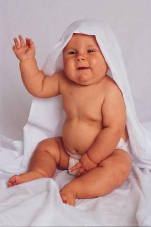 un bébé assis sur un tissu blanc, portant une serviette blanche sur la tête qui salue avec sa main